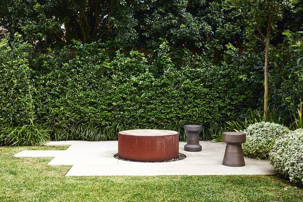 A landscaped outdoor garden seating area with a pale paved patio set into a lawn, surrounded by dense layered greenery. The space features a low round rust-coloured sculptural table or fire feature and two dark sculptural stools, creating a minimalist contemporary courtyard-like setting.