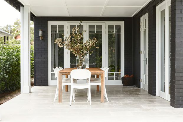 Covered outdoor dining terrace with a timber dining table and white chairs, set against a dark painted brick facade with white-framed glazed doors and windows. The space has a tiled floor, white ceiling, and a sheltered, leafy residential setting.