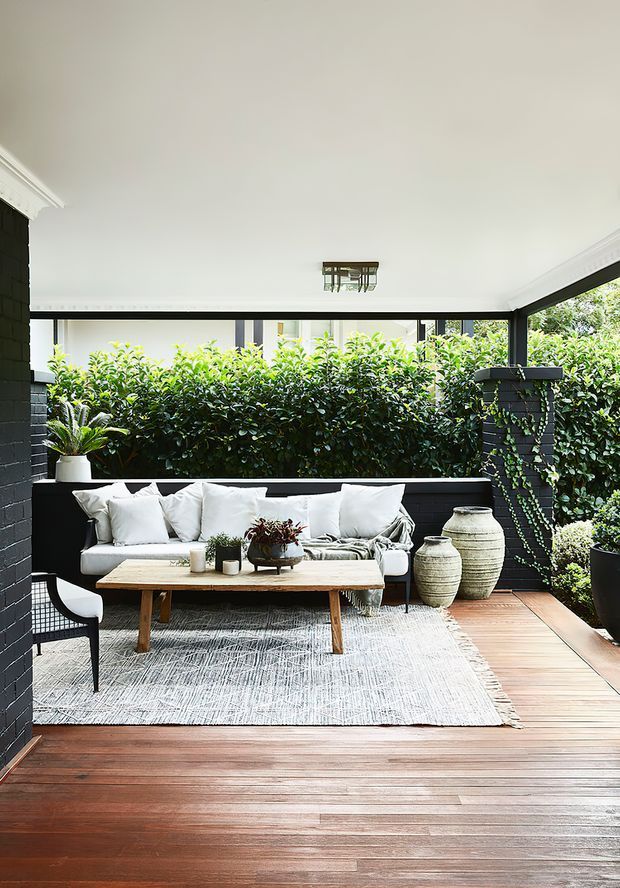 A bright, enclosed outdoor living terrace with a timber deck, black-painted masonry walls, extensive glazing, a pale upholstered sofa, coffee table, outdoor chairs, patterned rug, potted plants, and large decorative ceramic vessels. Dense greenery provides privacy beyond the terrace, while a simple ceiling light and dark-framed windows create a refined indoor-outdoor setting.