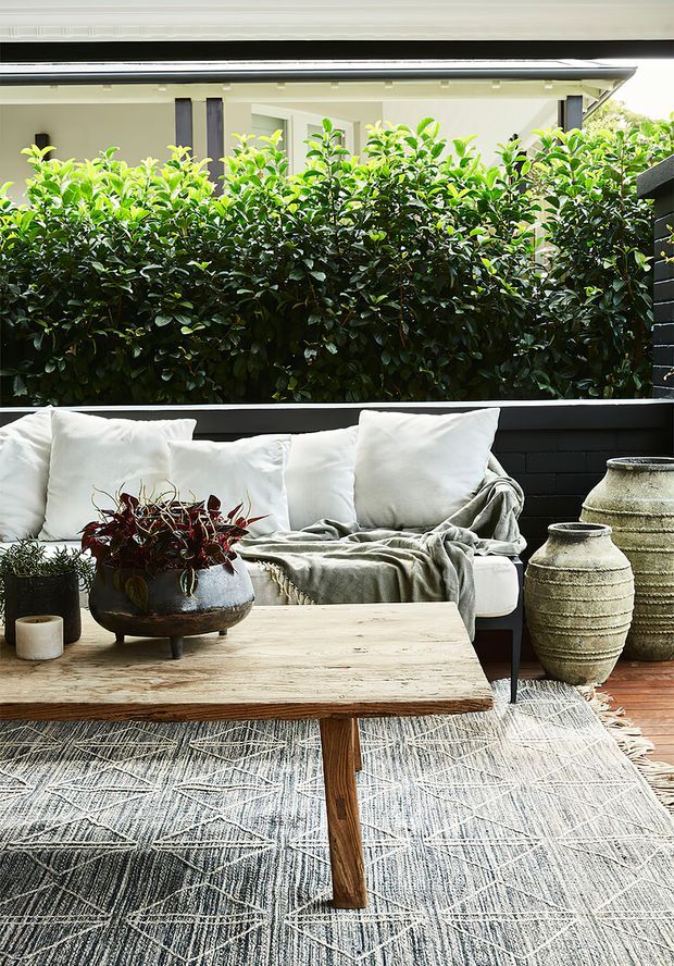 A bright, styled outdoor lounge area with a white cushioned sofa, grey throw, large patterned rug, rustic timber coffee table, ceramic vessels, and potted plants. Dense greenery forms a privacy screen behind a low black-painted masonry wall, with a modern roofed structure visible beyond.