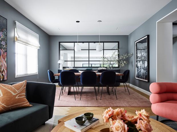 A refined open-plan dining and living area with dark grey walls, a large black-framed internal glass partition, and contemporary furniture. A dark dining table with navy upholstered chairs sits beneath spherical pendant lights, while a black sofa and sculptural coral-pink lounge chair frame the foreground. Layered rugs, artwork, a round timber coffee table, and floral decor add warmth and colour.