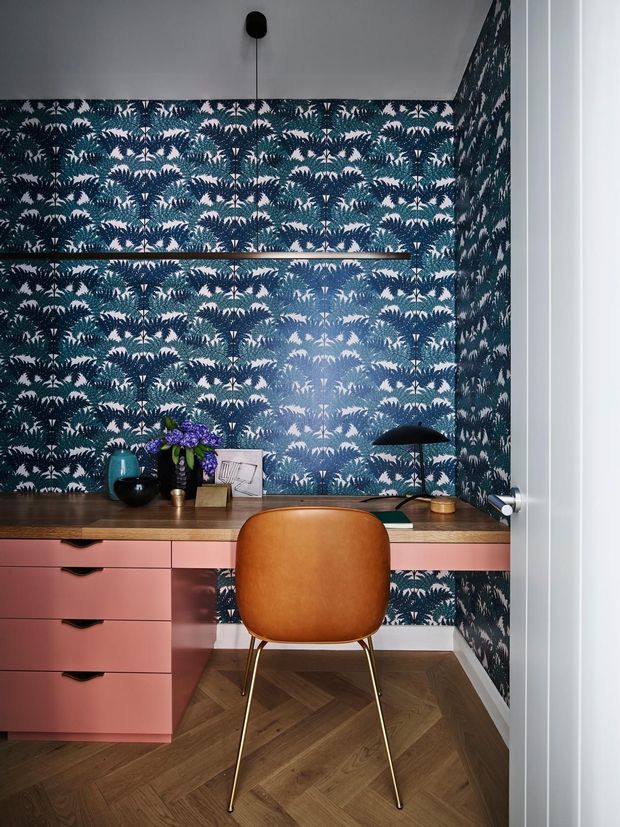 A compact, stylish home office nook viewed through an open doorway. The space features a wall-to-wall botanical patterned wallpaper in deep blue and teal, a built-in wooden desk with pink drawers, a tan upholstered chair with slim brass-toned legs, a black desk lamp, and decorative accessories. The floor is finished in dark herringbone timber, while the ceiling and door are white. The design has a bold, playful, retro-inspired character with a warm and inviting atmosphere.