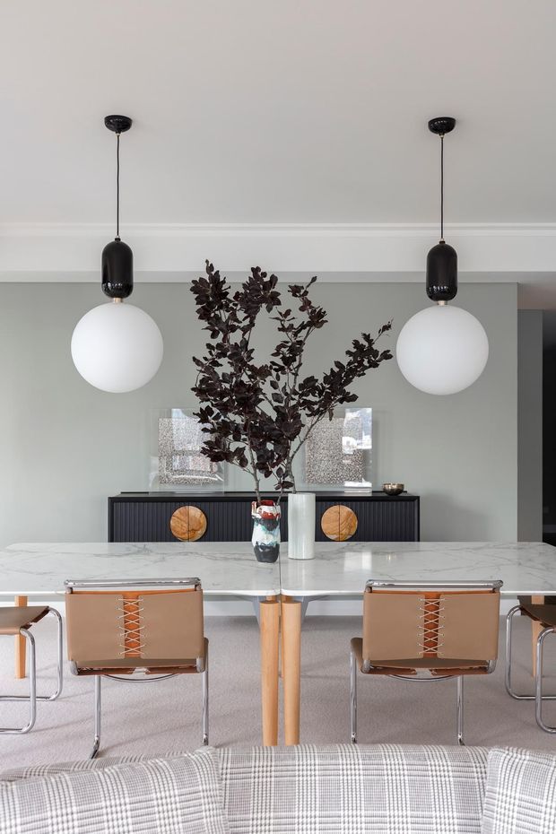 A refined dining area viewed from behind a sofa, featuring a long white marble dining table with tan leather and chrome-framed chairs, a dark fluted sideboard, sculptural pendant lights, and a large dark-leaf arrangement. The room has soft grey walls, white moulded ceiling trim, pale carpet, and a restrained, contemporary palette with warm wood and leather accents.