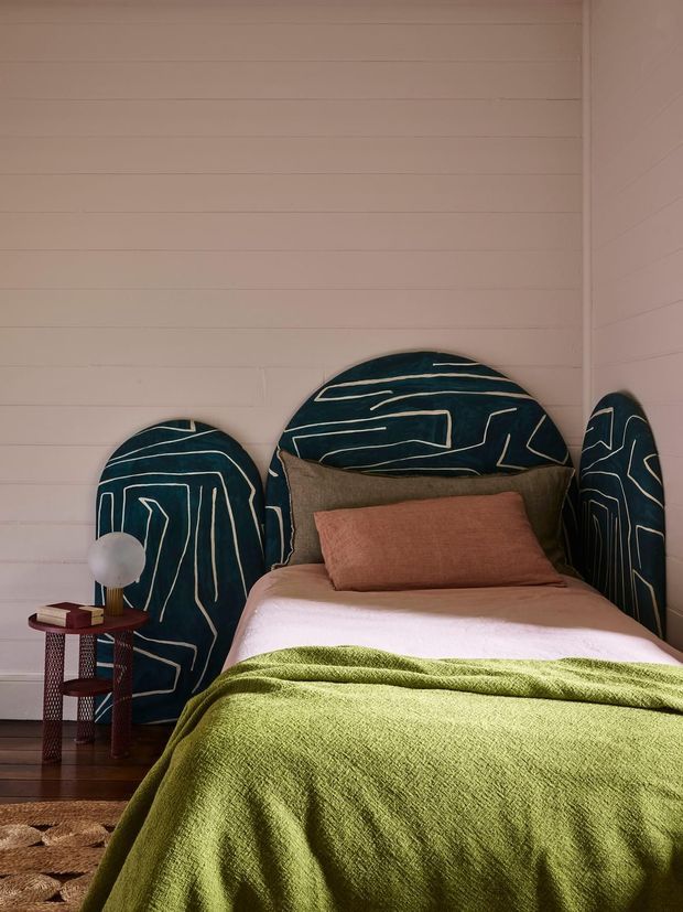 A cozy bedroom featuring a single bed with a sculptural dark teal upholstered headboard and matching side panels decorated with cream linear patterns. The bed is layered with white bedding, a rust-brown cushion, and a textured olive-green throw. White horizontal wall panelling forms the backdrop, with a small dark wood and red metal bedside table and spherical lamp beside the bed.