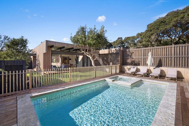 Exterior view of a contemporary timber-clad home with a large swimming pool and integrated shallow spa. The landscaped backyard includes timber decking, a timber privacy fence, lounge chairs, umbrellas, and a covered outdoor living area with glazing opening toward the pool.