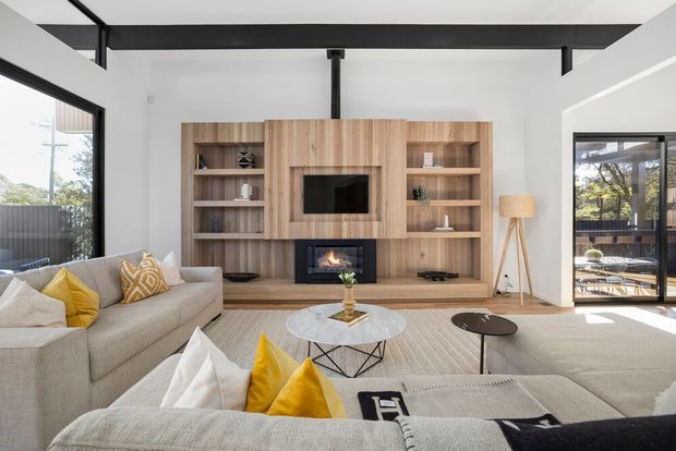 Bright contemporary living room with a large L-shaped light-grey sectional sofa, yellow and white cushions, a round coffee table, and a full-height timber built-in entertainment wall with open shelving, television niche, and fireplace. Black exposed steel beams contrast with white walls and ceiling, while large glazed openings bring in natural light and connect to an outdoor area.