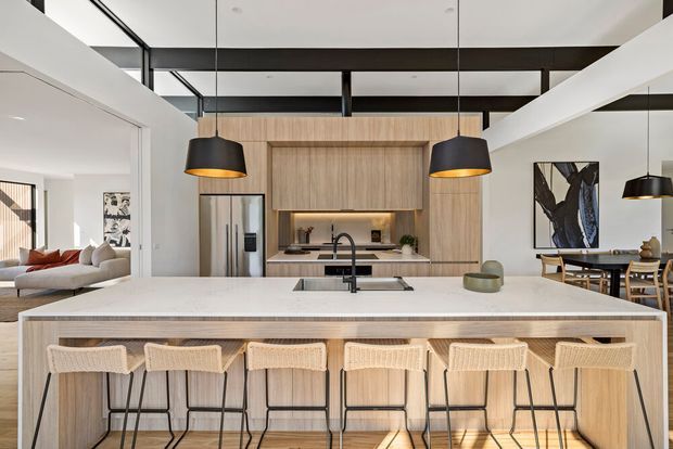 A bright, spacious contemporary kitchen with a large light-wood island and six woven bar stools, white stone-look surfaces, integrated timber cabinetry, a black gooseneck tap, stainless-steel refrigerator, and black pendant lights. Open-plan living and dining areas are visible beyond the kitchen, with exposed dark ceiling beams and extensive internal glazing creating a refined, airy feel.