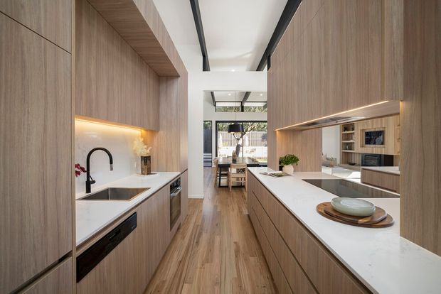 A contemporary galley-style kitchen with extensive light timber cabinetry, white stone benchtops, integrated appliances, black fixtures and framed glazing at the rear. A central island with seating faces a dining area, while concealed linear lighting and dark ceiling beams add contrast.