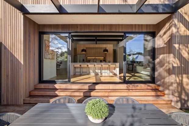 Exterior view of a contemporary timber-clad home opening onto a raised timber deck and outdoor dining area. Large black-framed glazed sliding doors reveal a bright open-plan kitchen with a central island, pendant lights, and bar stools. A projecting glazed canopy provides shelter above the entry, while strong sunlight creates patterned shadows across the facade and deck.