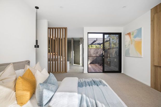 Bright contemporary bedroom with a neutral upholstered bed, layered white and blue bedding, and mustard accent cushions. A timber vertical-slat screen separates the sleeping area from the hallway, while black-framed glazing opens to a small timber deck or balcony. White walls, soft carpet, timber cabinetry, and a simple pendant light create a clean, relaxed interior.
