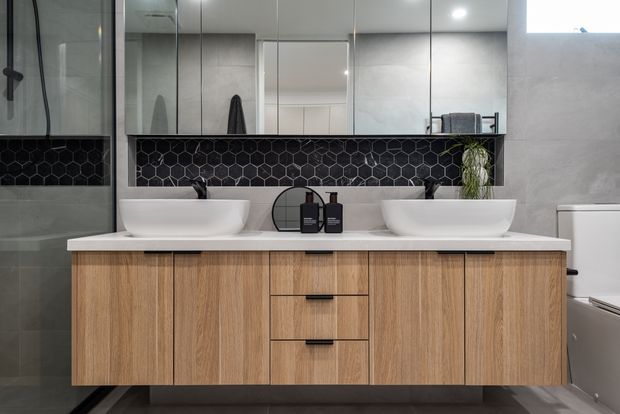 Contemporary double-vanity bathroom with a wall-mounted light timber vanity, white vessel basins, black tapware, a black hexagonal tile splashback, large mirrored cabinetry, and pale grey wall surfaces. A glass shower enclosure and freestanding bath are partially visible.