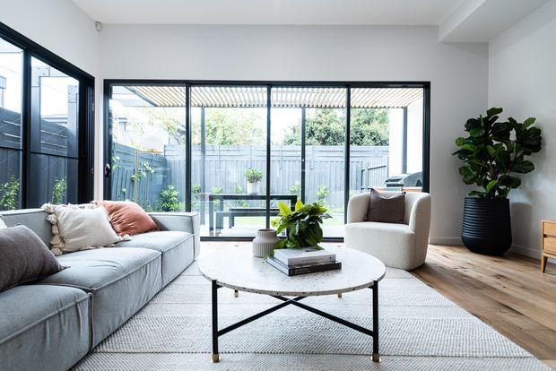 Bright contemporary living room with a large light-grey sectional sofa, round marble-look coffee table, textured rug, cream armchair, indoor plants, and warm timber flooring. Full-height black-framed glazing opens the room visually to a fenced garden courtyard.
