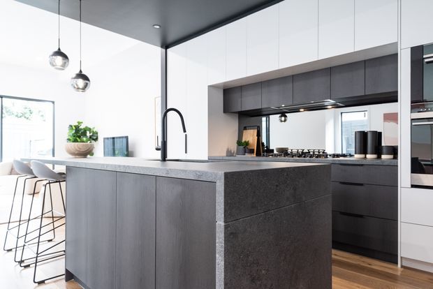 A contemporary open-plan kitchen dominated by a large dark grey stone island with integrated seating and a minimalist black gooseneck tap. Full-height white cabinetry, dark lower cabinetry, a concealed rangehood, integrated appliances, pendant lights and timber flooring create a refined, high-contrast interior. A dining area and large window are visible beyond the island.
