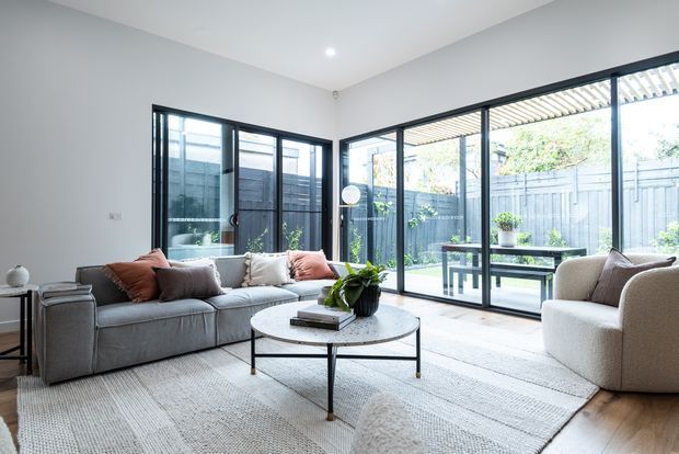 Bright contemporary living room with a large grey sectional sofa, round coffee table, textured area rug, and curved upholstered armchair. Full-height black-framed glazing opens visually onto a paved courtyard, with a pergola and garden planting outside. The neutral interior is softly accented with rust and mauve cushions and greenery.
