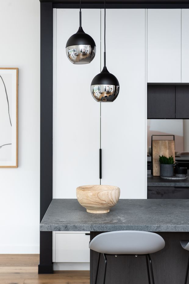 A refined contemporary kitchen interior featuring a dark charcoal stone island benchtop with a light timber bowl, dark wood island cabinetry, white flat-panel cabinetry, and two black-and-chrome pendant lights. A grey upholstered bar stool sits at the island, with black-framed glazing or cabinetry and pale timber flooring adding contrast. The image emphasizes the island, pendant lighting, and clean monochromatic material palette.