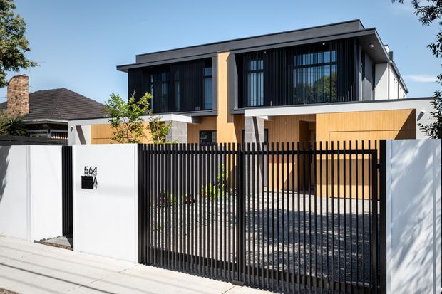 Street-facing view of a contemporary two-storey residence with a flat roof, dark vertical window screens, warm timber-toned cladding, white rendered walls, and a black vertical-slat security fence and gate. The front entry and paved driveway are visible, with a neighbouring older home partially visible behind.