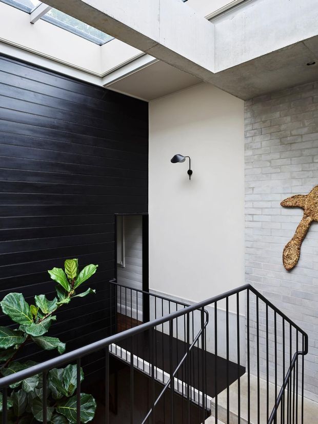 Modern interior stairwell or landing with black-painted horizontal wall cladding, pale rendered walls, exposed concrete beams, skylights, and a light brick feature wall. A black metal balustrade surrounds the staircase, with indoor greenery and sculptural wall art adding visual interest.