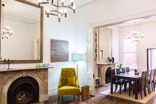 Bright traditional living and dining interior with ornate fireplaces, elaborate mouldings, large decorative mirrors, timber flooring, and a contemporary geometric pendant light. A mustard-yellow tufted armchair adds a bold accent beside the living-room fireplace, while the adjoining dining room contains a dark timber dining table and chairs, another fireplace, and tall windows.