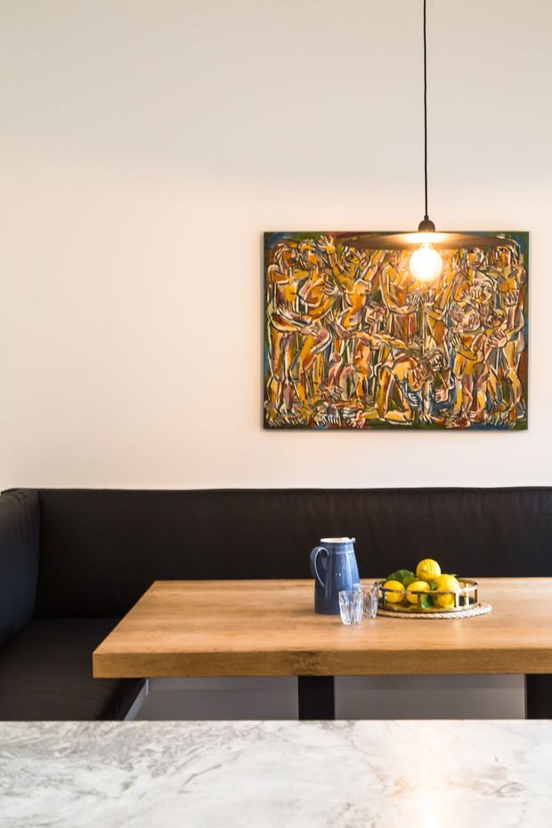 A contemporary dining nook with a built-in black upholstered banquette, a thick timber dining table, and a white marble or stone foreground countertop. A colourful abstract artwork hangs on the pale wall beneath a simple black pendant light. A blue jug, drinking glasses, and a bowl of citrus sit on the table.