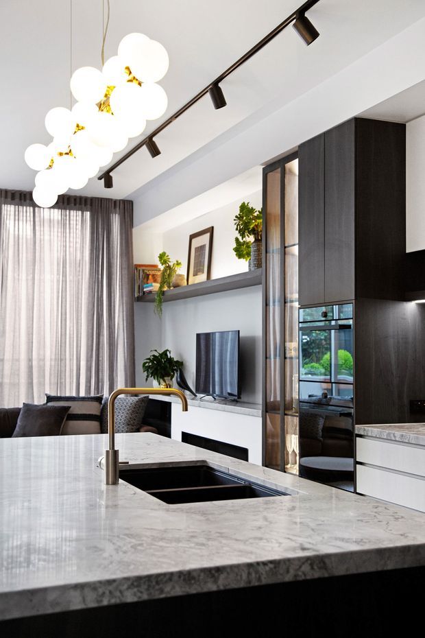 A contemporary open-plan kitchen viewed from behind a large stone island with an undermount sink and brass-toned tap. The kitchen features dark vertical-grain cabinetry, integrated black appliances, pale stone benchtops, glass-fronted joinery, and a white living area beyond with a television, shelving, plants, and layered pendant and track lighting.