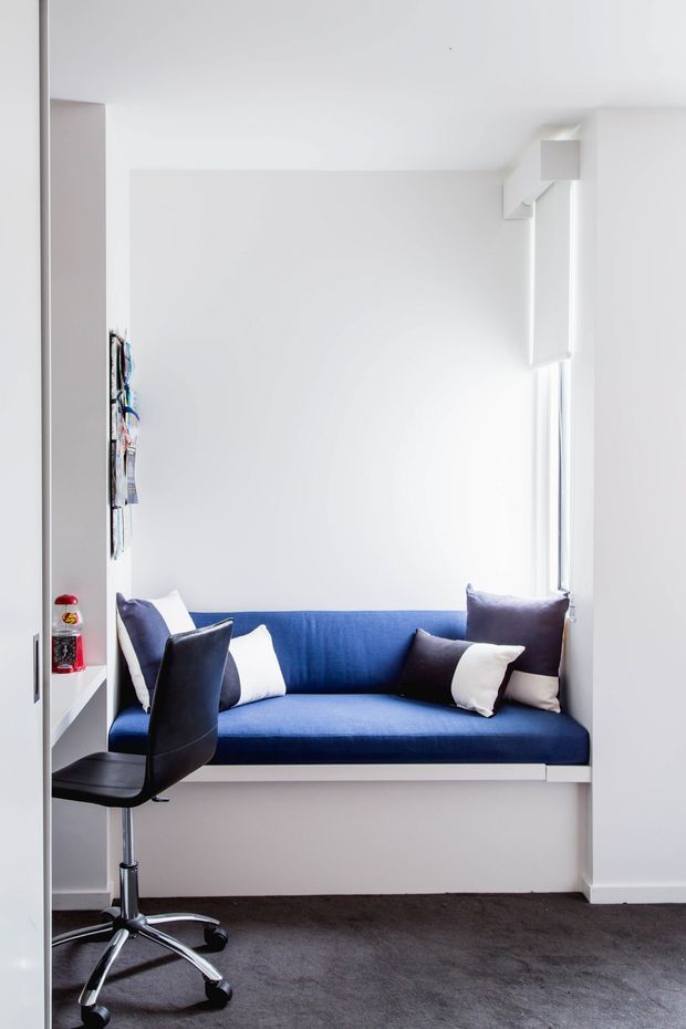 Bright, compact white interior nook with a built-in deep blue window seat, contrasting cushions, a black swivel office chair, and dark carpet. Minimal built-in joinery and a tall narrow window create a clean, functional workspace or reading area.