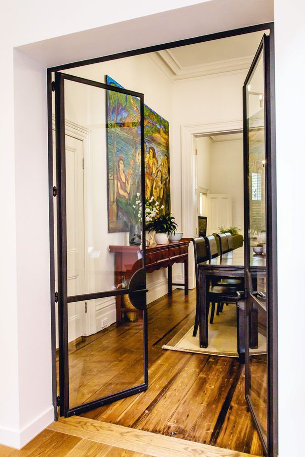 A bright, elegant dining area viewed through open black metal-framed glazed doors. The room features polished warm timber flooring, white walls with decorative trim and moulding, a wooden console table, colourful artwork, and a dark dining table with upholstered chairs. The glazed doors create strong reflections and a sense of visual connection between adjoining rooms.