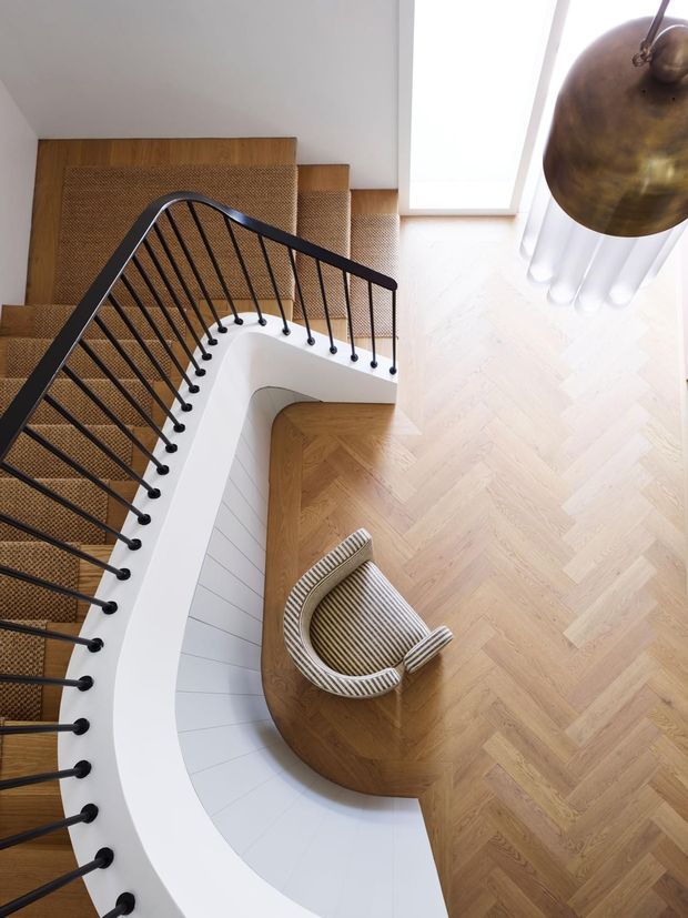 A top-down view of a curved interior stairway with a white sculptural balustrade and dark metal spindle railing. The stairs are carpeted in a warm natural-fibre tone, while the surrounding floor is laid in light herringbone timber. A curved timber landing includes a striped upholstered chair, and a large bronze pendant hangs beside tall sheer curtains and a bright window.