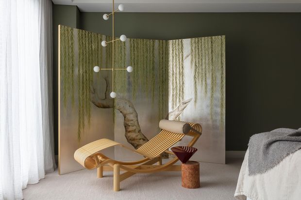 A sophisticated bedroom interior with dark olive-green walls, sheer white curtains, pale carpet, and a sculptural folding screen featuring a botanical tree image. A curved wooden chaise lounge and matching ottoman sit in front of the screen, complemented by a modern globe pendant light and a partially visible bed with layered grey and white bedding.