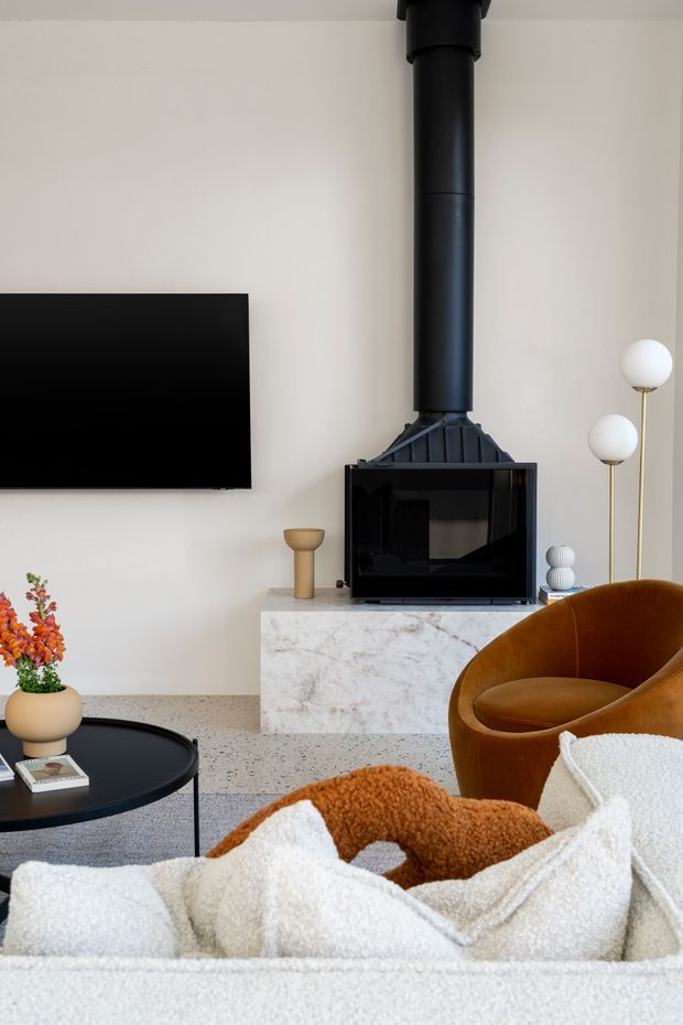 A contemporary living room featuring a black freestanding wood-burning fireplace with a tall flue, mounted flat-screen television, pale walls, and a white marble-clad hearth or media cabinet. The room includes a sculptural brown leather lounge chair, a black round coffee table, a light upholstered sofa with textured throws, and decorative lighting and accessories. The overall palette is warm neutral with black accents and a refined, cozy atmosphere.