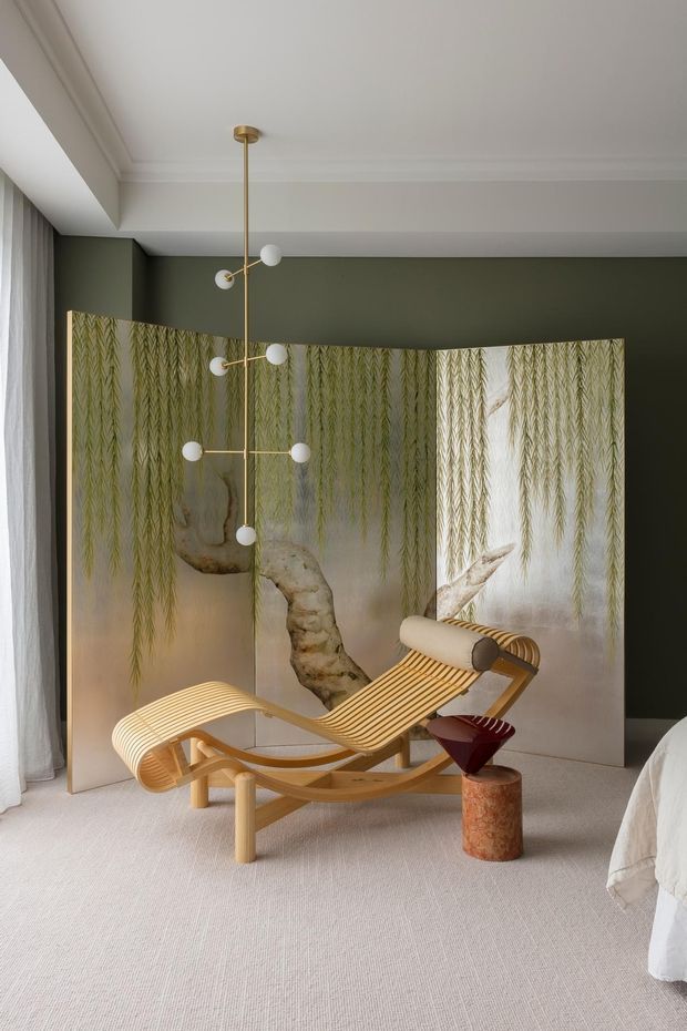 A refined bedroom interior with a sculptural wooden chaise lounge, a small side table, and a contemporary brass-and-opal globe pendant light. The room features a deep olive-green accent wall, white decorative ceiling mouldings, sheer curtains, pale carpet, and a large botanical screen or artwork behind the seating. The edge of a bed is visible at right, confirming the bedroom setting. The palette is warm, muted, and sophisticated with natural timber and botanical imagery.