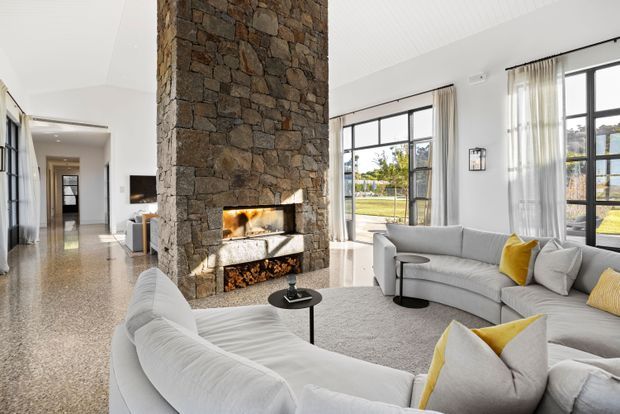 Bright, spacious contemporary living room with a large curved white sectional sofa, yellow accent cushions, expansive black-framed windows, polished terrazzo flooring, and a dramatic full-height natural stone fireplace feature. A hallway and additional interior spaces are visible beyond the living area.