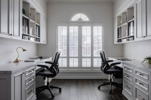 Bright, symmetrical home office with two built-in workstations facing each other, white paneled cabinetry and open shelving, dark office chairs, and a large central window with plantation shutters and a fanlight. The room has a refined, traditional feel with dark timber flooring and brass-accented task lighting and hardware.