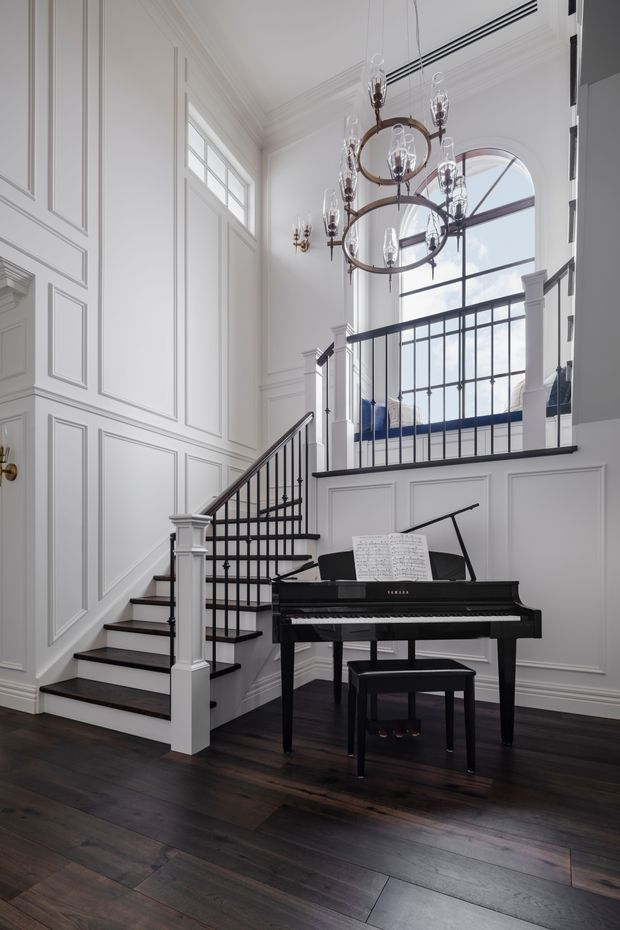 A refined, double-height stair hall with a black grand piano in the foreground. The space features dark timber flooring, white decorative wall panelling, a black metal balustrade, a sculptural circular chandelier, and a large arched window overlooking an upper-level landing.
