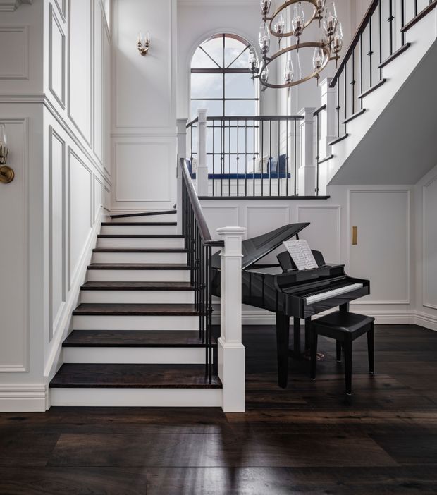 A refined residential stair hall featuring a dark timber staircase and flooring, white decorative wall panelling, black metal balustrades, and a glossy black grand piano beneath a multi-tiered chandelier. A tall arched window brings daylight into the double-height circulation space, with a landing and upper-level railing visible beyond.