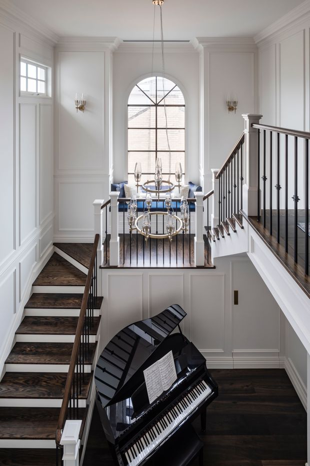 A refined, light-filled multi-level stair hall with a central landing and dining or sitting area beyond. White decorative wall panelling, classical mouldings, dark timber flooring, black metal balustrades, and a large arched window create a formal traditional interior. A glossy black grand piano occupies the lower level, while ring chandeliers and wall sconces add elegant detailing.