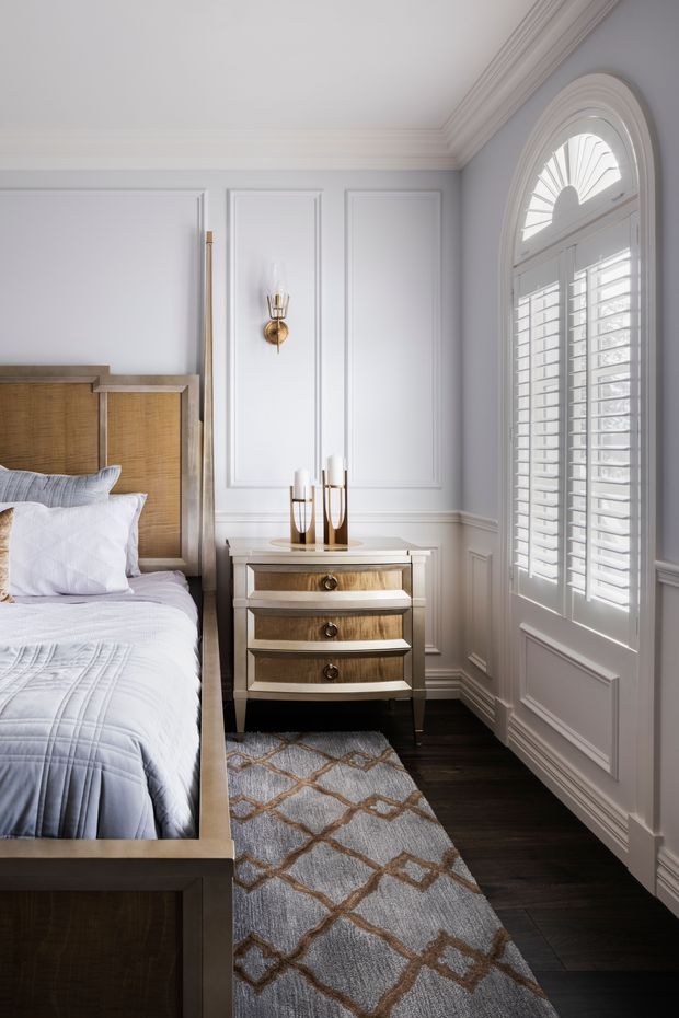 A refined bedroom corner with a partially visible upholstered bed, warm wood headboard, and a small three-drawer bedside chest. The room features pale grey-white walls with decorative raised wall panelling, ornate crown moulding and skirting, dark timber flooring, and a tall arched window fitted with white plantation shutters. Soft blue-grey bedding and a patterned runner rug create a calm, classic interior.