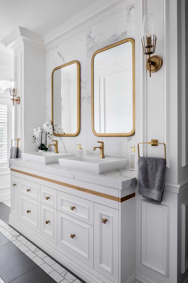 A refined double-sink bathroom vanity with white framed cabinetry, a white marble-look benchtop and splashback, gold tapware and hardware, rounded rectangular gold-framed mirrors, and decorative wall sconces. Traditional wall mouldings and panelled cabinetry are paired with crisp white finishes and charcoal floor tiles.