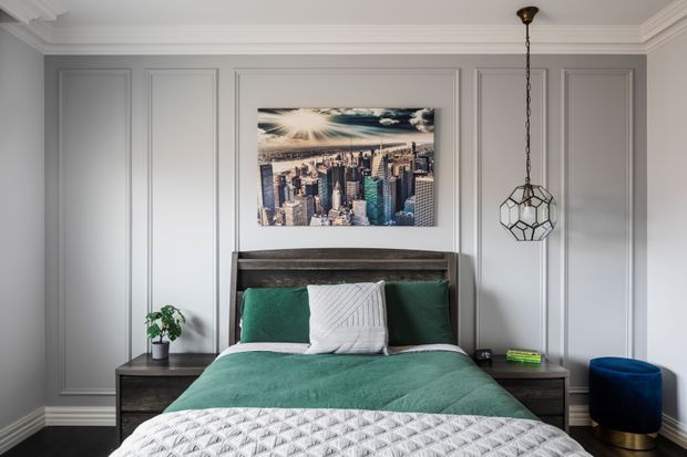 A refined bedroom with a centered bed, grey decorative wall panelling, a dark timber headboard and bedside cabinetry, and a green bedspread with layered cushions. A framed cityscape artwork hangs above the bed, while a geometric pendant light and small blue accent chair add visual interest. The room has detailed ceiling cornices, trim, and skirting boards.