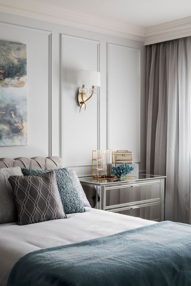 Elegant bedroom with a soft grey upholstered bed, pale bedding, layered blue-grey throw and cushions, mirrored bedside chest, sculptural gold wall sconce, full-height grey curtains, and light grey walls featuring decorative raised-panel moulding. The room has a refined, calm, contemporary-classic aesthetic with cool neutral tones and blue accents.