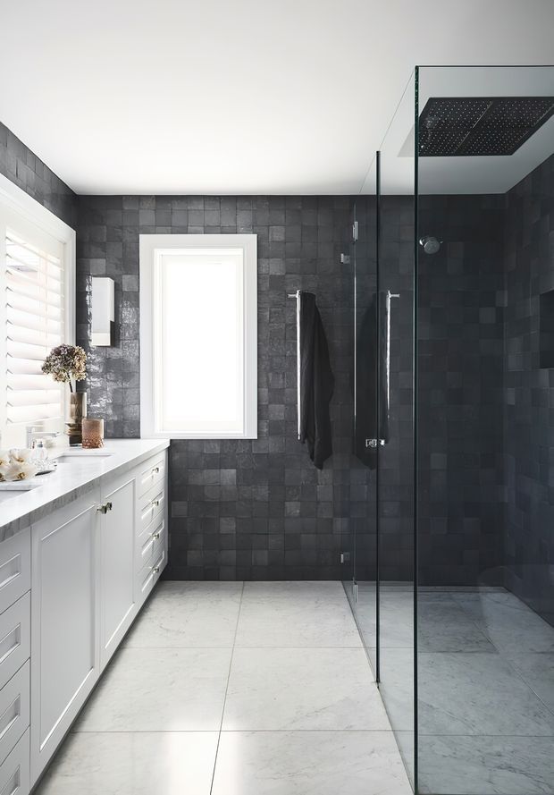 A bright contemporary bathroom with a long white vanity and marble-look floor tiles. The walls and walk-in shower are finished in dark charcoal square tiles, with frameless glass shower panels, chrome fixtures, a large rainfall showerhead, and white plantation shutters. The room has a clean, high-contrast monochromatic scheme.