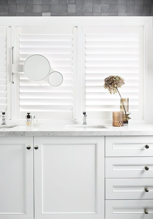 Bright, refined bathroom vanity with white shuttered windows, a pale marble benchtop, white framed cabinetry, chrome tapware, and understated decorative accessories. The composition is clean and predominantly white, with a grey stone tile band above the windows.