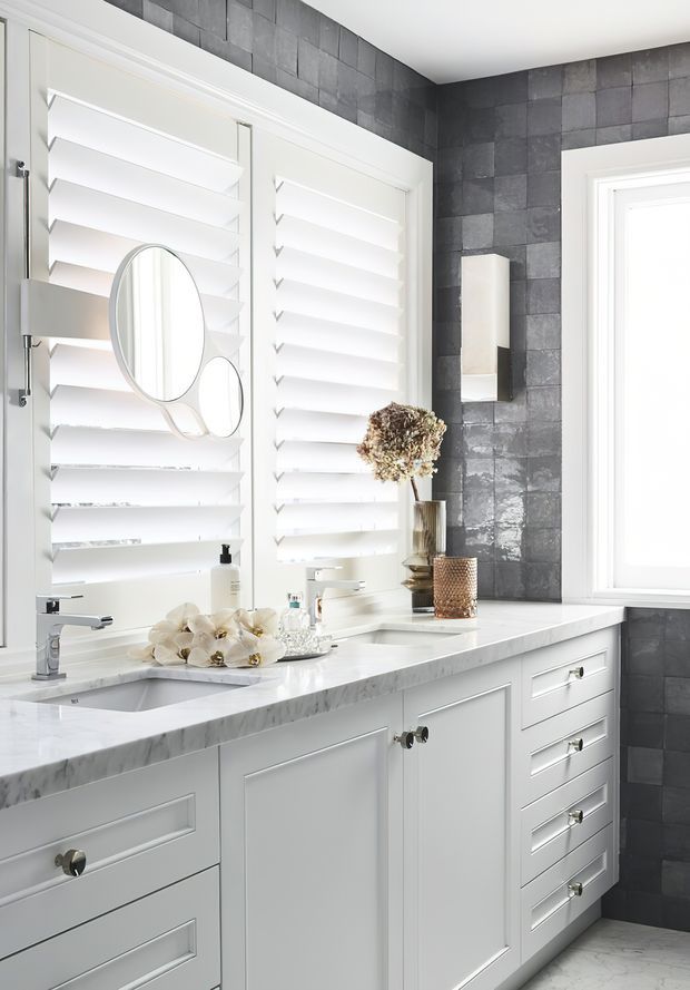 Bright, refined bathroom vanity with extensive white shaker-style cabinetry, a marble-look countertop and integrated basin. Dark grey square wall tiles create a strong contrasting backdrop, while white plantation shutters, framed mirrors, chrome hardware and decorative accessories complete the space.