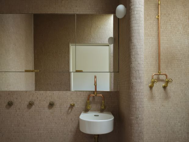 Compact bathroom with a wall-mounted white basin, exposed brass plumbing and taps, and a large mirrored cabinet. The walls and cylindrical column are covered in small beige mosaic tiles, creating a warm, tactile, refined interior. Minimal spherical wall lights and a partially visible frosted window add to the understated design.