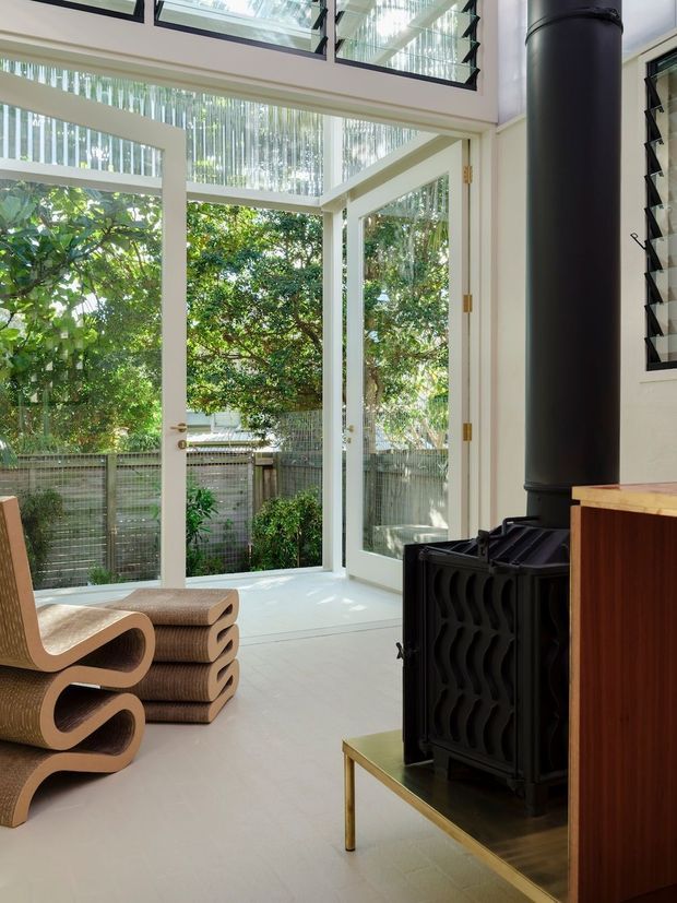 Bright contemporary living space with floor-to-ceiling glazing and louvred windows opening onto a leafy outdoor area. A black freestanding wood-burning stove sits beside a warm timber cabinet, while sculptural corrugated cardboard lounge chairs add a distinctive feature. The room has a light neutral palette, abundant natural light, and a strong connection to the garden.