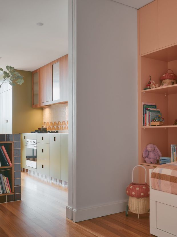 A warm, colourfully styled interior showing a child’s bedroom in the foreground with a built-in coral-pink shelving unit, books, toys, a bed and a small woven bedside basket. Through the opening is a compact kitchen with pale cabinetry, timber wall cupboards, patterned tiled splashback and a muted yellow-green wall. Timber flooring runs through both spaces, with white trim and soft natural light.