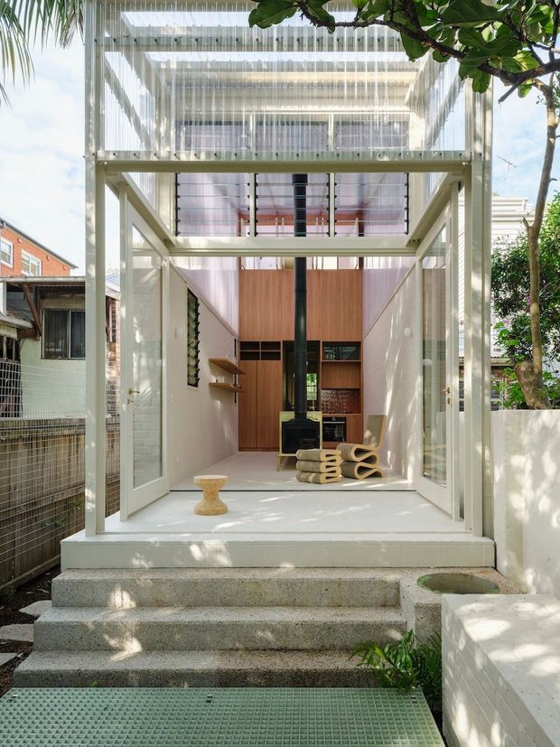 A bright, narrow contemporary house extension or entry pavilion viewed from the garden. The space has a highly glazed, open framed structure with translucent corrugated or ribbed screening overhead and across the upper level, white-painted structural framing, white tiled or rendered walls, and concrete steps leading into the interior. Warm timber wall panelling and built-in cabinetry are visible at the rear, along with a black central flue or stove and sculptural neutral-toned seating. Mature greenery surrounds the pavilion, while neighbouring residential buildings are visible at the sides.