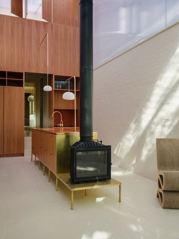 A refined open-plan interior combining a kitchen and living area. Warm timber wall panelling and built-in cabinetry surround a compact kitchen island with a reddish stone benchtop and brass-toned base. A freestanding black wood-burning stove with a tall flue sits on a brass-toned platform, while sculptural pendant lights, pale flooring, a high white wall, and a textured woven lounge chair complete the space. Strong natural light creates dappled shadows across the wall and floor.