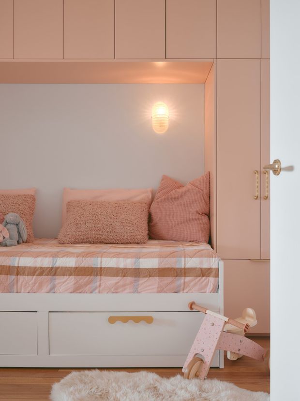 A compact, built-in children's bedroom nook with a raised bed surrounded by dusty pink cabinetry and panelling. The bed has a plaid pink-and-beige quilt, textured pink cushions, and a small plush toy. A warm wall sconce illuminates the pale grey-blue back wall, while a pink scooter rests on a fluffy rug over timber flooring. The space feels playful, cosy, and carefully integrated into the joinery.