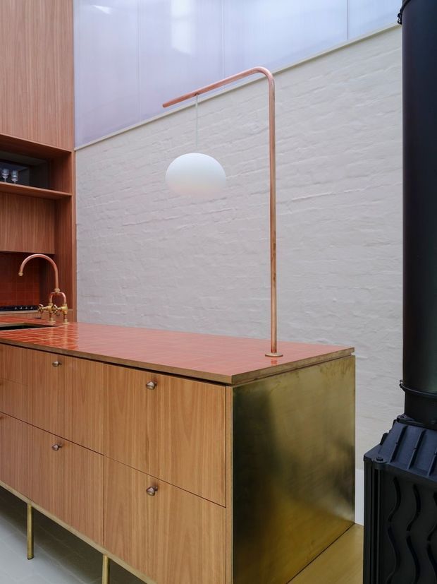 A close-up view of a refined kitchen with warm timber cabinetry, a terracotta-red tiled benchtop and splashback, a brass-toned end panel, white-painted brick walls, and a slender copper floor lamp. A black freestanding wood-burning stove is visible at the right edge. The space combines tactile materials and warm metallic details with a clean, contemporary composition.