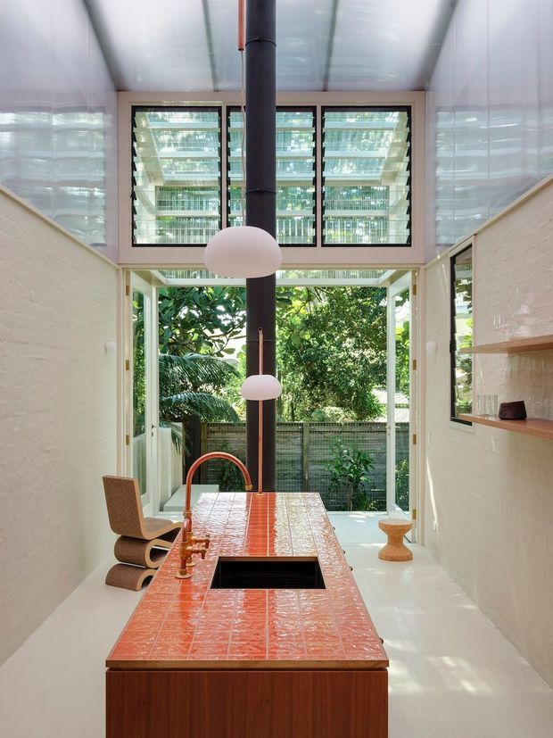 A narrow, light-filled kitchen with a long red-orange tiled island and integrated black sink, copper-toned tapware, white-painted brick walls, and a dark cylindrical flue descending through the centre. Large black-framed louvred windows and glazed doors open visually toward a leafy courtyard. White pendant lights, timber cabinetry, open wall shelving, and polished light flooring complete the contemporary space.